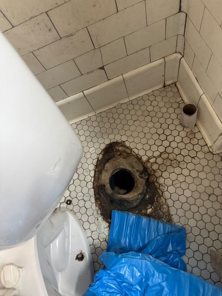 Old toilet removed exposing the cast iron floor flange and wax ring, with blue sheeting protecting the tile floor