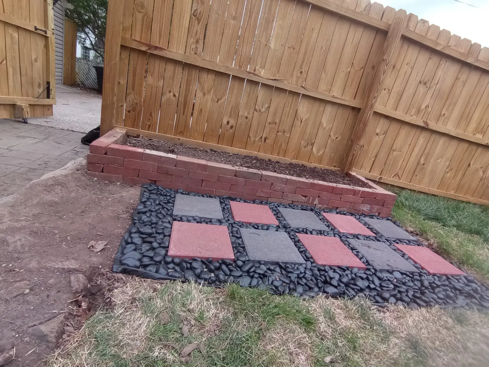 Raised brick planter bed with red and grey paver stepping stones set in black river rock beside a timber fence