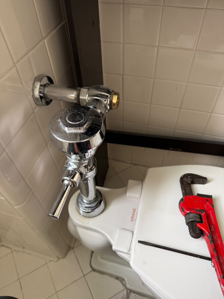 Chrome flushometer flush valve being serviced with a pipe wrench resting on the toilet tank lid